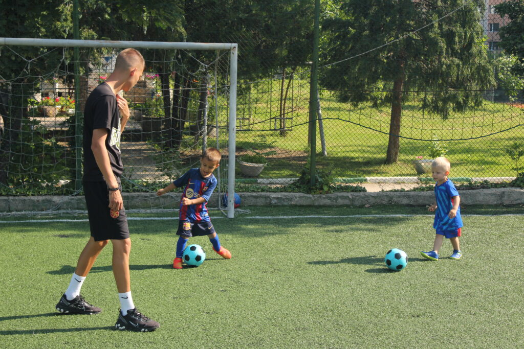 Две деца ритат топка. Тренировка по футбол за деца 3-6 години. Треньора коментира играта.